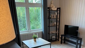 Una sala de estar con sofá, una mesa de centro, un televisor sobre un mueble, una estantería y una ventana con cortinas.