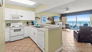 Cocina con gabinetes blancos, microondas y estufa. Hay un fregadero y una pequeña isla con una cafetera. La habitación tiene un balcón con vistas al océano.