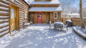 Una cabaña de troncos con una puerta roja, un porche cubierto con mobiliario exterior y un paisaje cubierto de nieve.