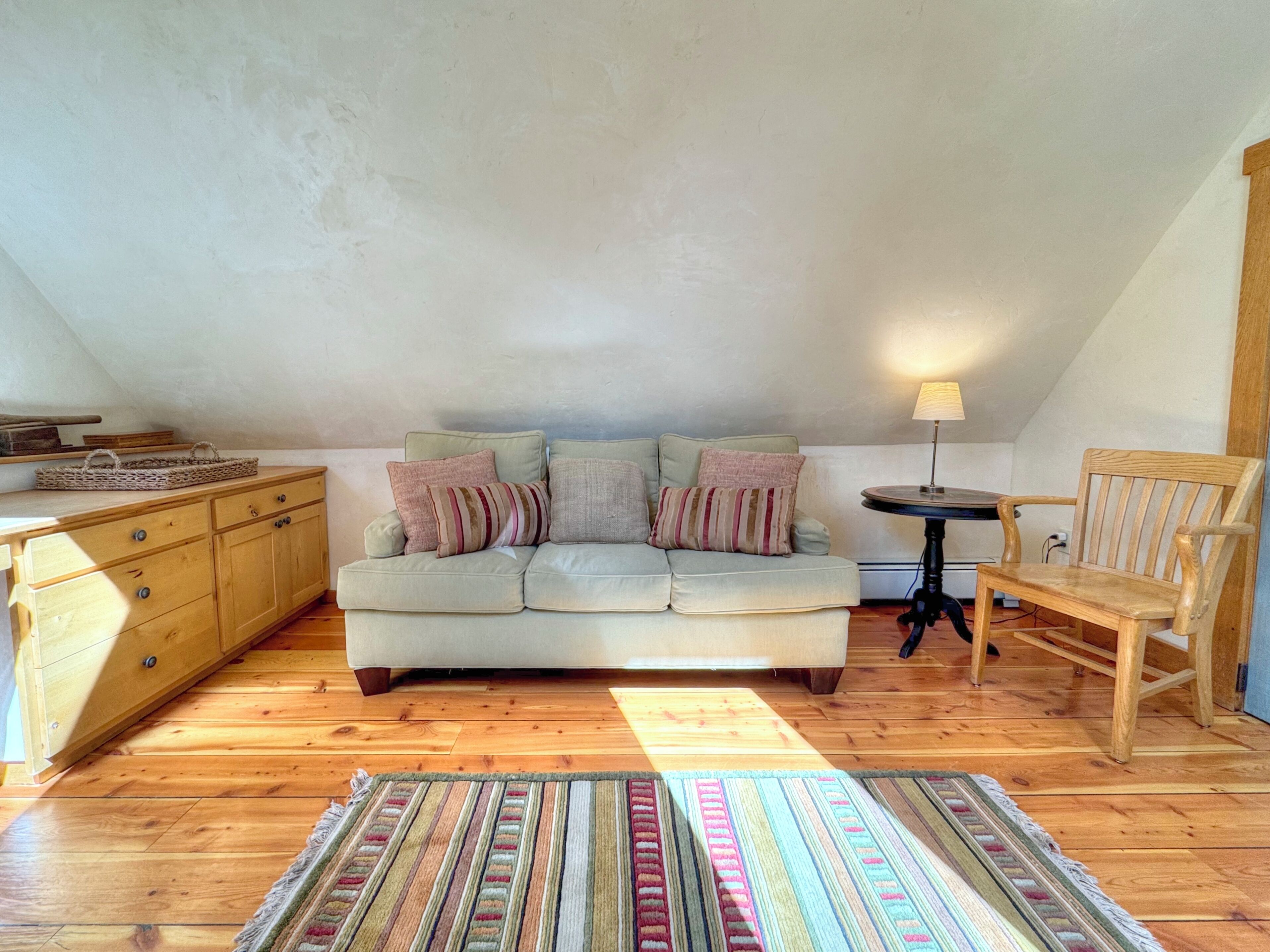 A cozy loft bedroom with a light-colored sofa, a wooden chair, a small table, and a patterned rug.