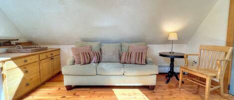 A cozy loft bedroom with a light-colored sofa, a wooden chair, a small table, and a patterned rug.