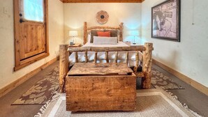 Un dormitorio rústico con estructura de cama de madera, una mesita de noche, una mesilla auxiliar, un adorno de pared y un cofre de madera grande.