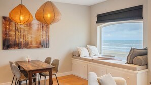 A dining area with a wooden table and chairs, a bench with cushions, and a view of the ocean through a large window. - Sandhues Beachfront Apartment (Zandvoort)