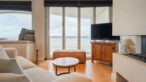 A modern living room with a sofa, a round table, a television, and a view of the sea through large windows. - Sandhues Beachfront Apartment (Zandvoort)