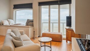 A living room with a sofa, ottoman, and a television on a wooden cabinet. A large window offers a view of the ocean. - Sandhues Beachfront Apartment (Zandvoort)