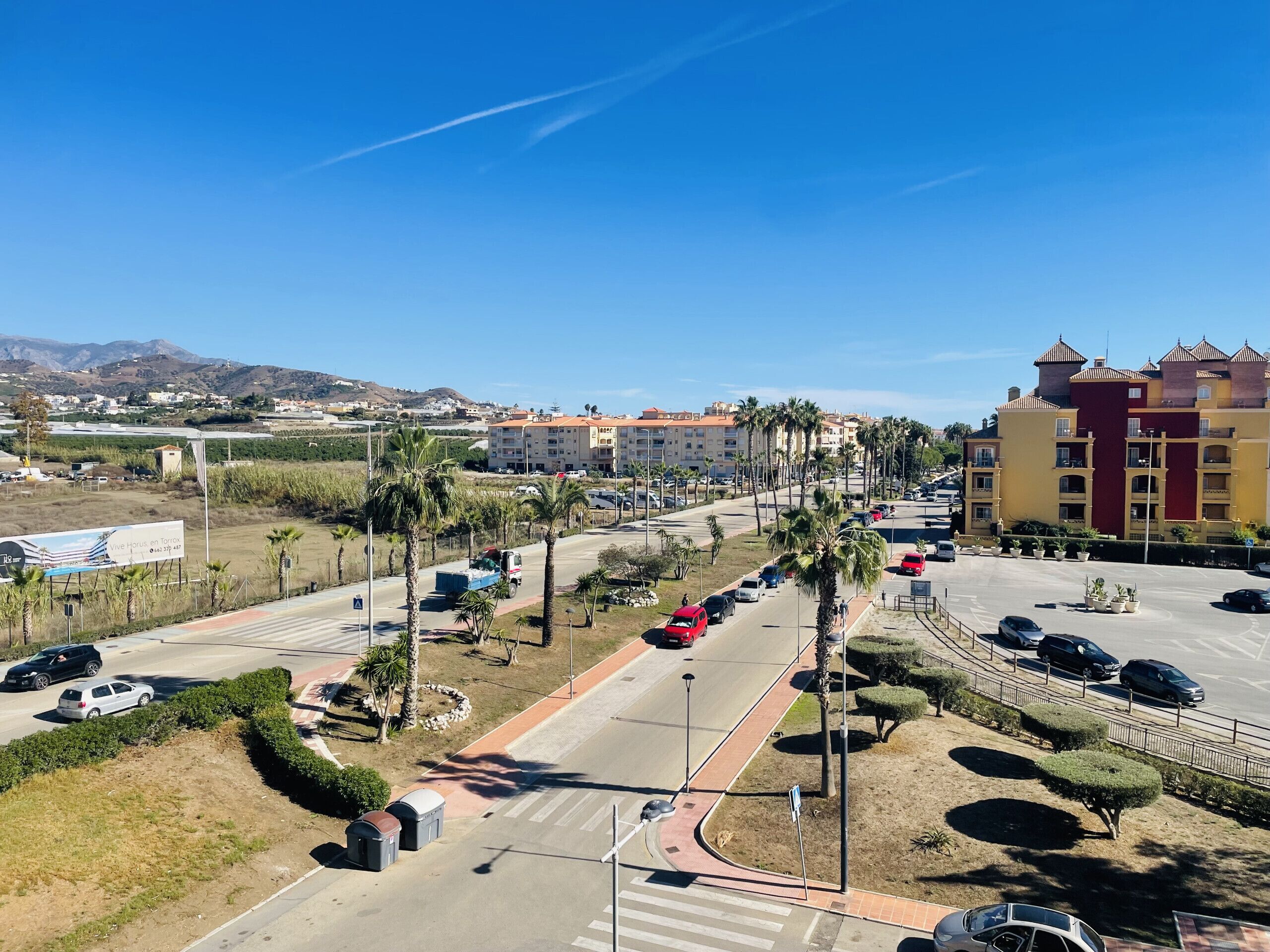 A parking lot with multiple cars, palm trees, and a building with a red and yellow facade.