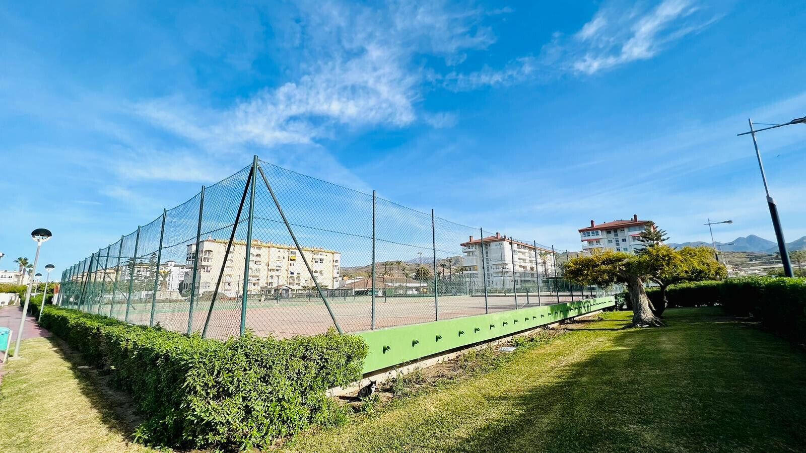 A tennis court enclosed by a green fence, surrounded by well-maintained grass and bushes, with buildings in the background.