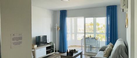 Ein modernes Wohnzimmer mit Sofa, Couchtisch, Fernseher und einem Balkon mit blauen Vorhängen.