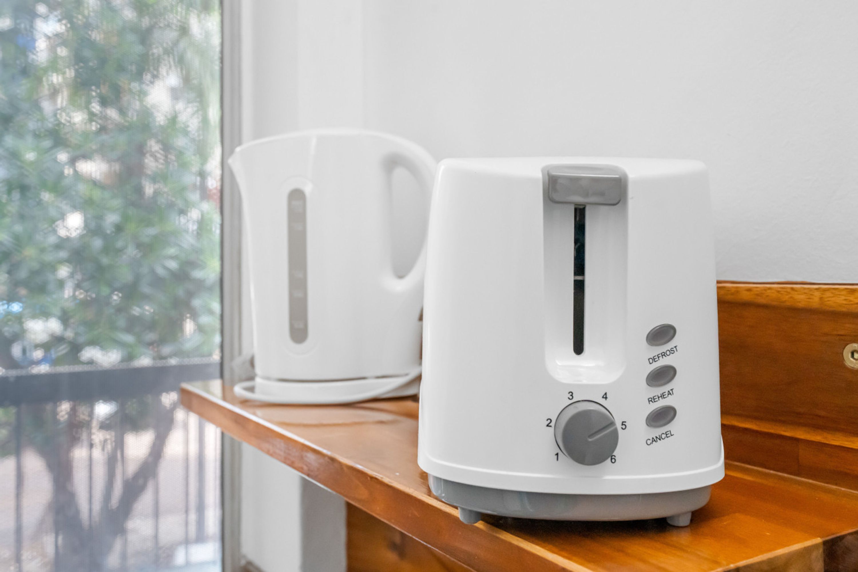 A white toaster on a wooden countertop next to a white mug, with a window in the background.