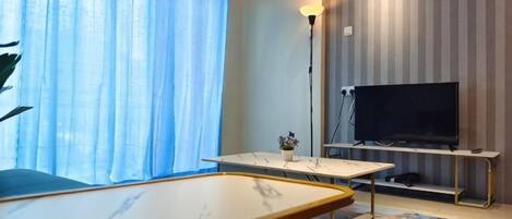 Ein Zimmer mit einem Flachbildfernseher auf einem Schrank, einem Tisch mit einer Lampe und einem Fenster mit Vorhängen.