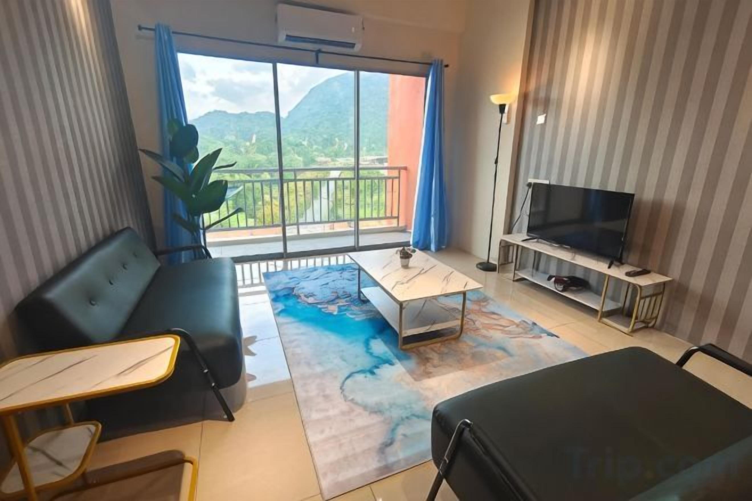 A modern hotel room with a balcony, a flat-screen TV, a sofa, a dining table, and a large window offering a mountain view.