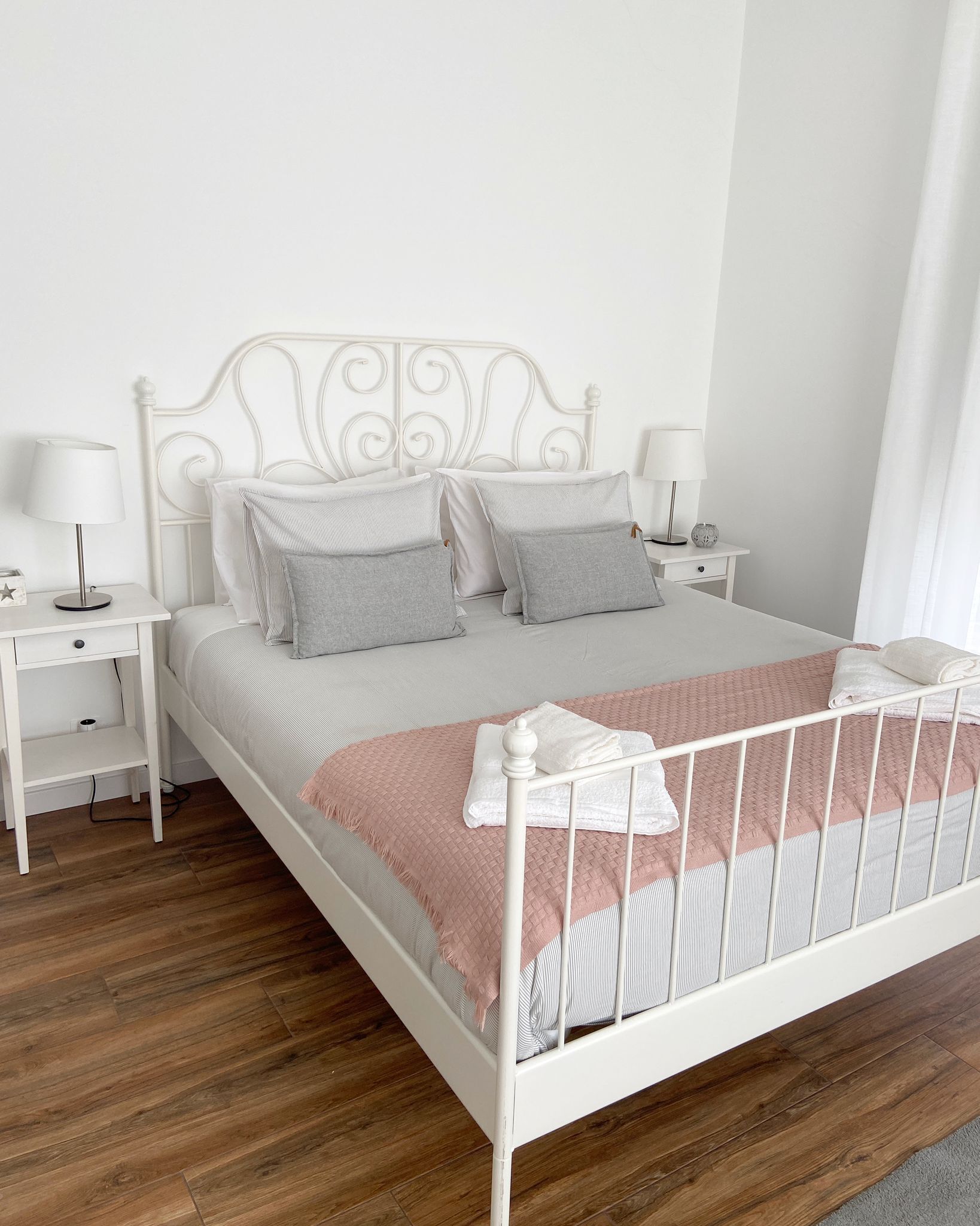 Uma cama de metal branca com colcha rosa, duas mesas de cabeceira com abajures e piso de madeira.