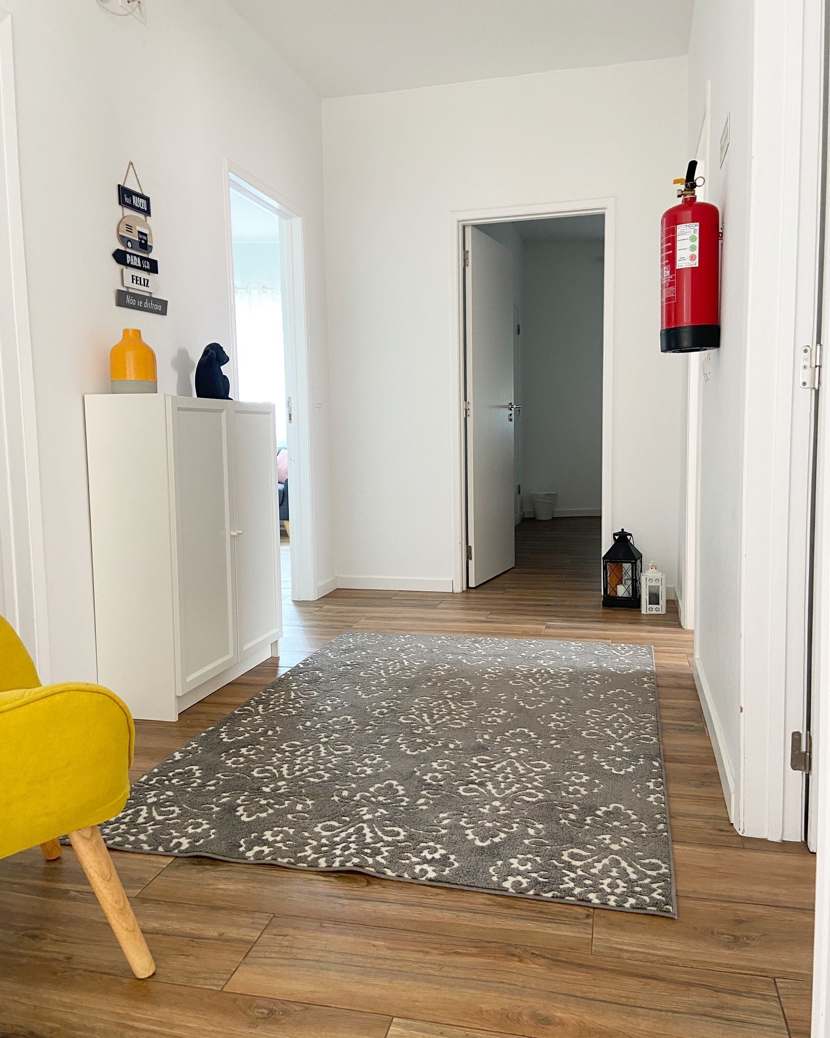 Un couloir avec un tapis à motifs, une armoire blanche, une chaise jaune et un extincteur.
