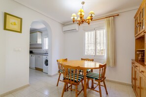 A dining area with a round wooden table, six chairs with striped cushions, a chandelier, an air conditioning unit, a window with curtains, and a kitchen visible through an archway.