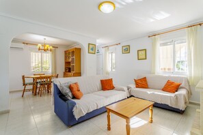 A living room with a sofa, a dining table, and chairs. There is a wooden cabinet and a chandelier.