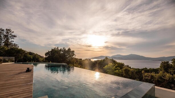Ein Pool im Infinity-Design mit Holzterrasse, umgeben von üppiger Vegetation und Blick auf das Meer und die Berge.
