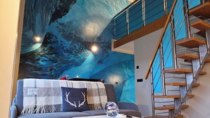 Un salon moderne avec un canapé gris, une table basse blanche et une fresque représentant une scène de forêt. On aperçoit, à l’arrière-plan, un escalier en bois avec des rampes en verre.