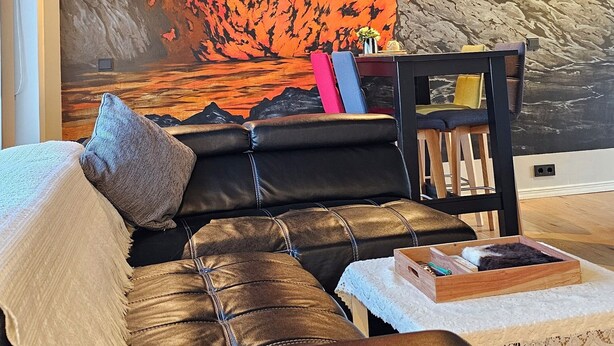 Un salon moderne agrémenté d’une grande fresque représentant une éruption volcanique, d’un canapé en cuir et d’une table basse en bois sur laquelle sont posés des livres.
