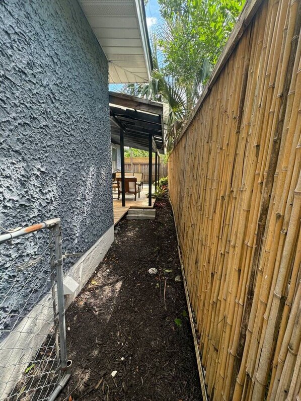 Et smalt udendørs område med en overdækket patio, bambushegn og en mørk, struktureret væg.