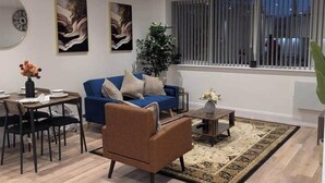 Una sala de estar moderna con un sofá azul, un sillón marrón, una mesa de comedor con sillas y un ventanal con persianas.