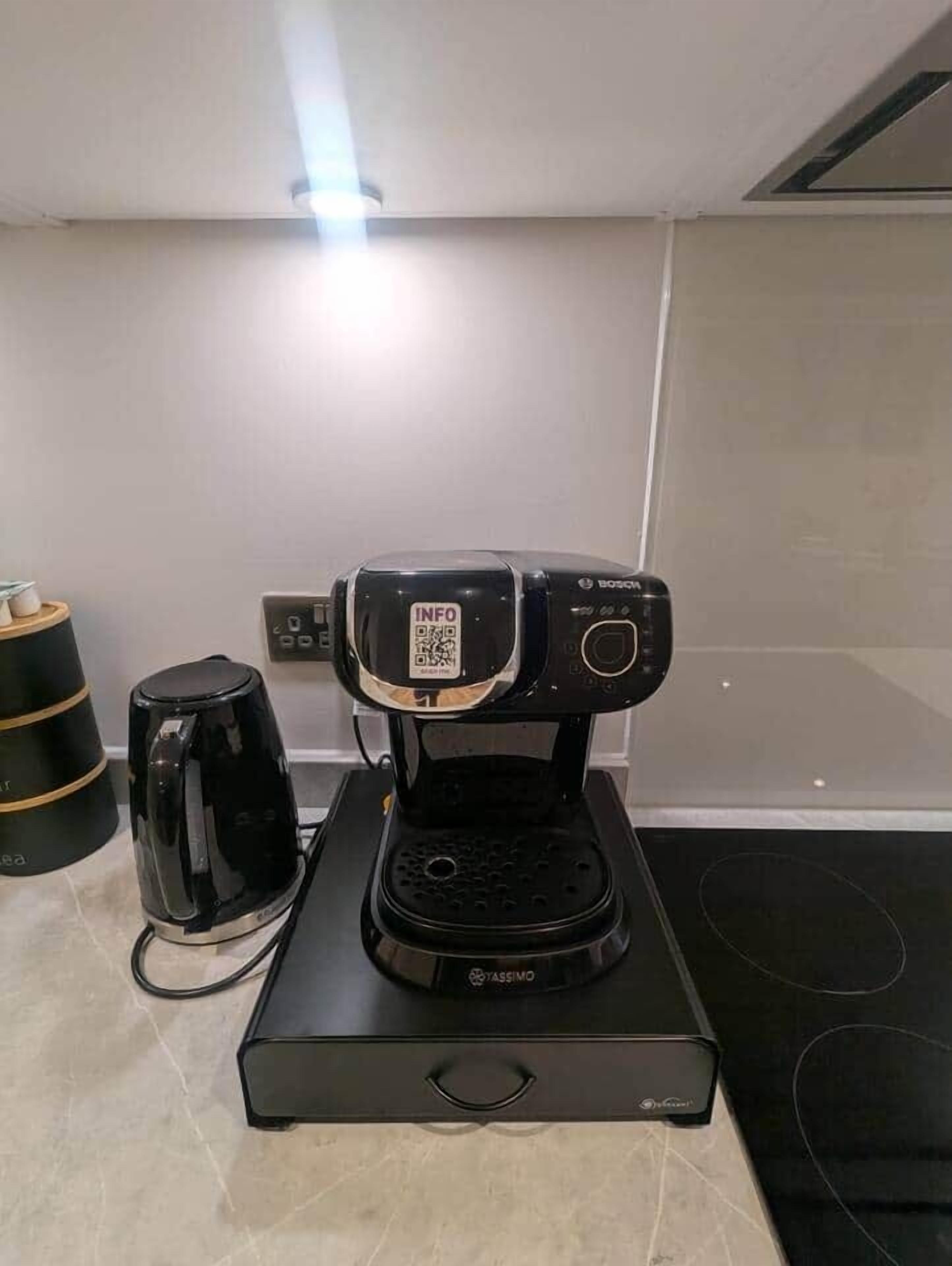 Una cafetera compacta sobre una base negra, una tetera eléctrica negra y una pequeña pila de tazas sobre una mesa.