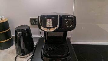 Una cafetera compacta sobre una base negra, una tetera eléctrica negra y una pequeña pila de tazas sobre una mesa.