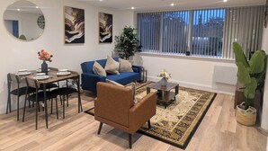 Una sala moderna con un sofá azul, un sillón marrón, una mesa de comedor con sillas y un amplio ventanal con persianas.