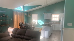 Uma sala de estar ampla com um sofá marrom, uma área de refeições com mesa e cadeiras, uma cozinha com armários brancos e uma janela grande que permite muita luz natural.