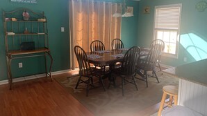 Uma sala de jantar com uma mesa redonda, seis cadeiras, um candelabro, uma janela com cortinas, uma estante e um aviso na parede.