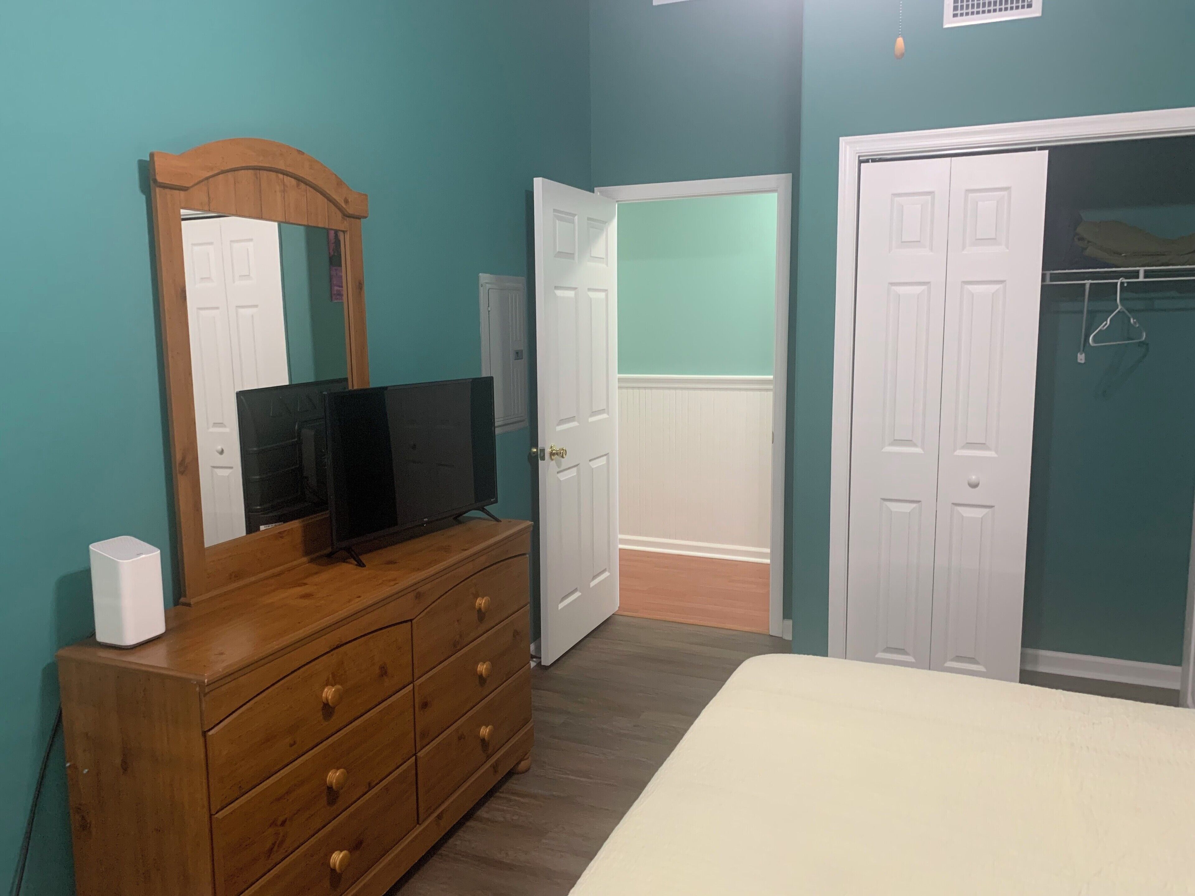Une chambre avec des murs de couleur turquoise, une commode en bois, un téléviseur et un lit blanc.
