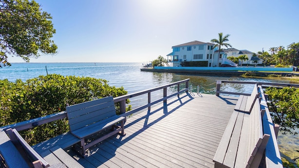 A wooden deck with benches overlooking a body of water, a white building with a balcony, and palm trees in the background. - Completely Updated 2BR Condo–Cedars East #613 by RVA (Longboat Key)