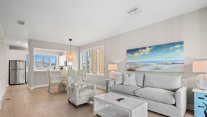 A modern living room with a grey sofa, a coffee table, and a patterned armchair. There is a dining area with a table and chairs, and a kitchen with a refrigerator and cabinets. - Completely Updated 2BR Condo–Cedars East #613 by RVA (Longboat Key)