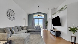 En moderne stue med en hjørnesofa, et fladskærms-tv, en loftventilator og et stort vindue med gardiner.