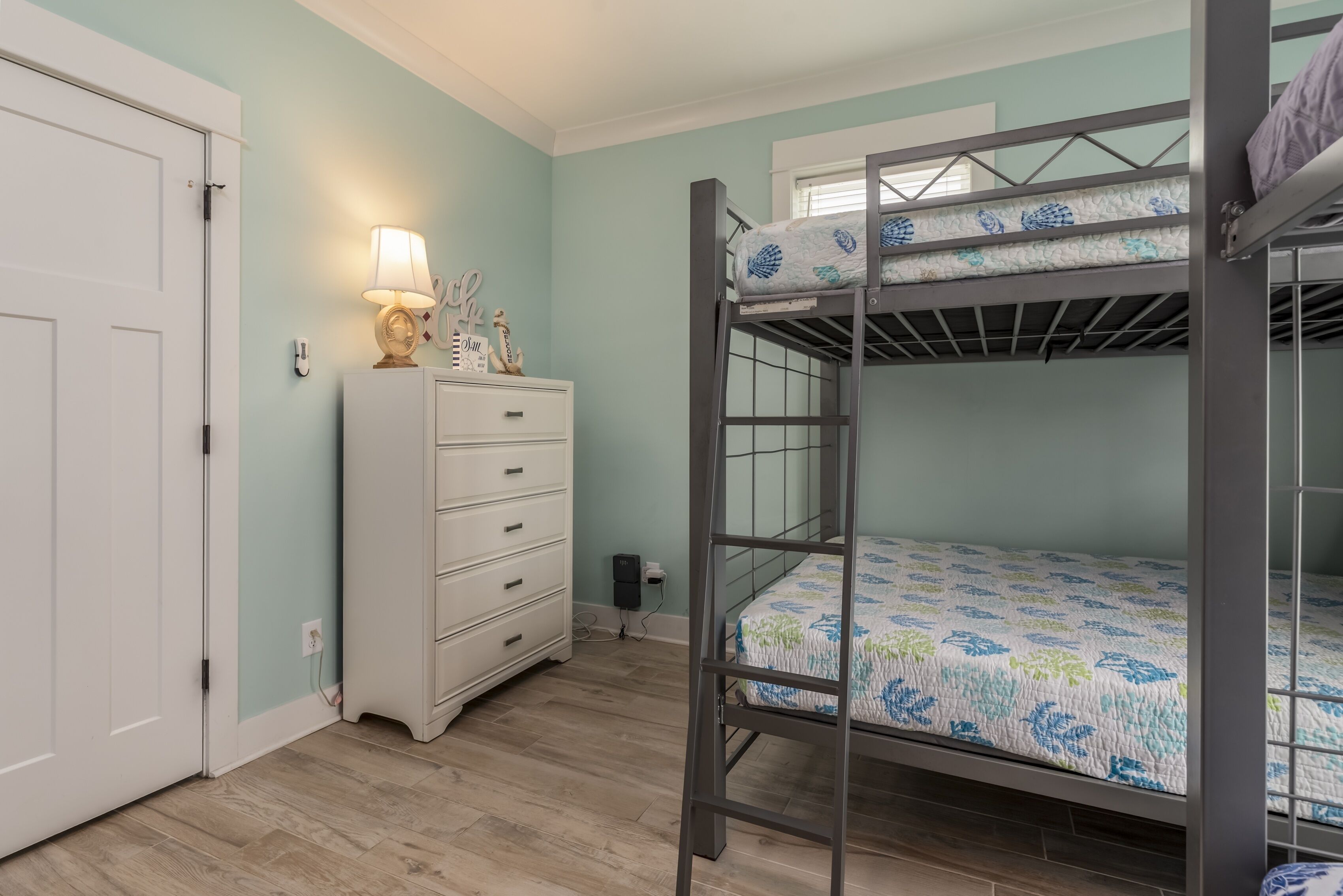 A bunk bed with a floral bedspread, a white dresser with drawers, a lamp, and a door.