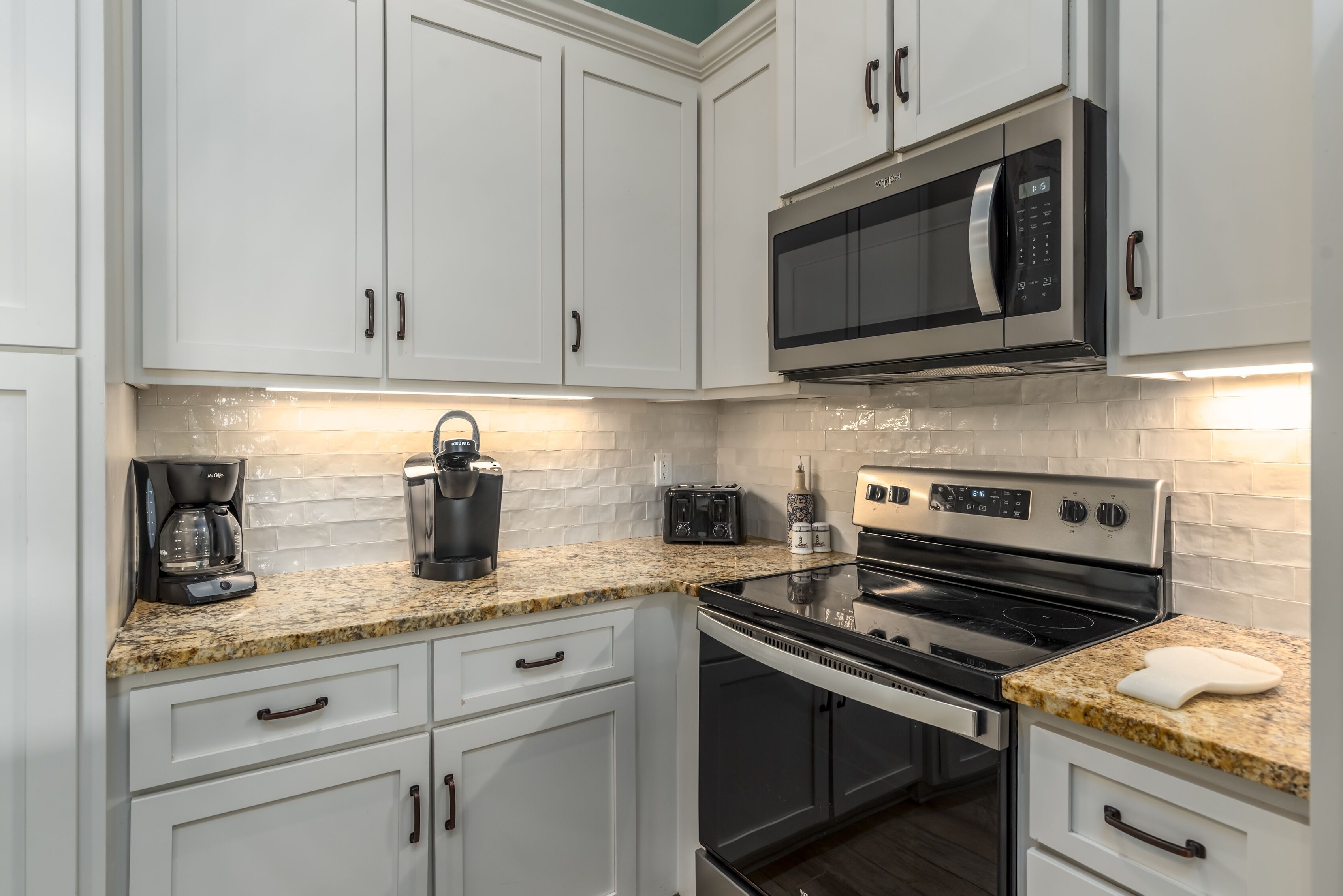 A modern kitchen with white cabinets, a granite countertop, a built-in microwave, a stove, and a coffee maker.