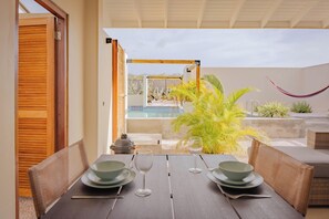 An outdoor dining area with a table set for two, a hammock, and a view of a pool and garden. - Bunitesa di bida Kas Iquana (Soto)