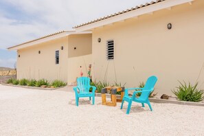 Un bâtiment de couleur beige, avec un toit recouvert de tuiles, deux chaises bleues, une petite table et une statue de flamant rose décorative.