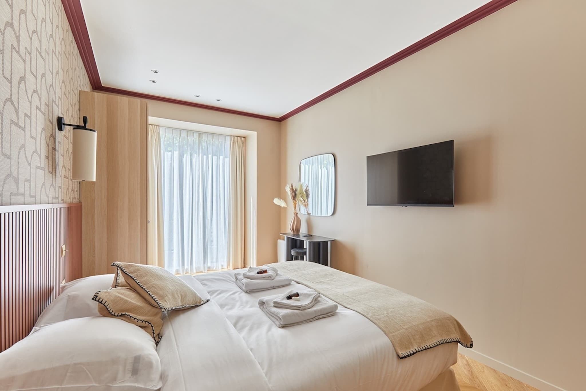 Une chambre d’hôtel avec un grand lit, un téléviseur à écran plat fixé au mur, une table de chevet ornée d’un vase de fleurs et une fenêtre avec des rideaux.