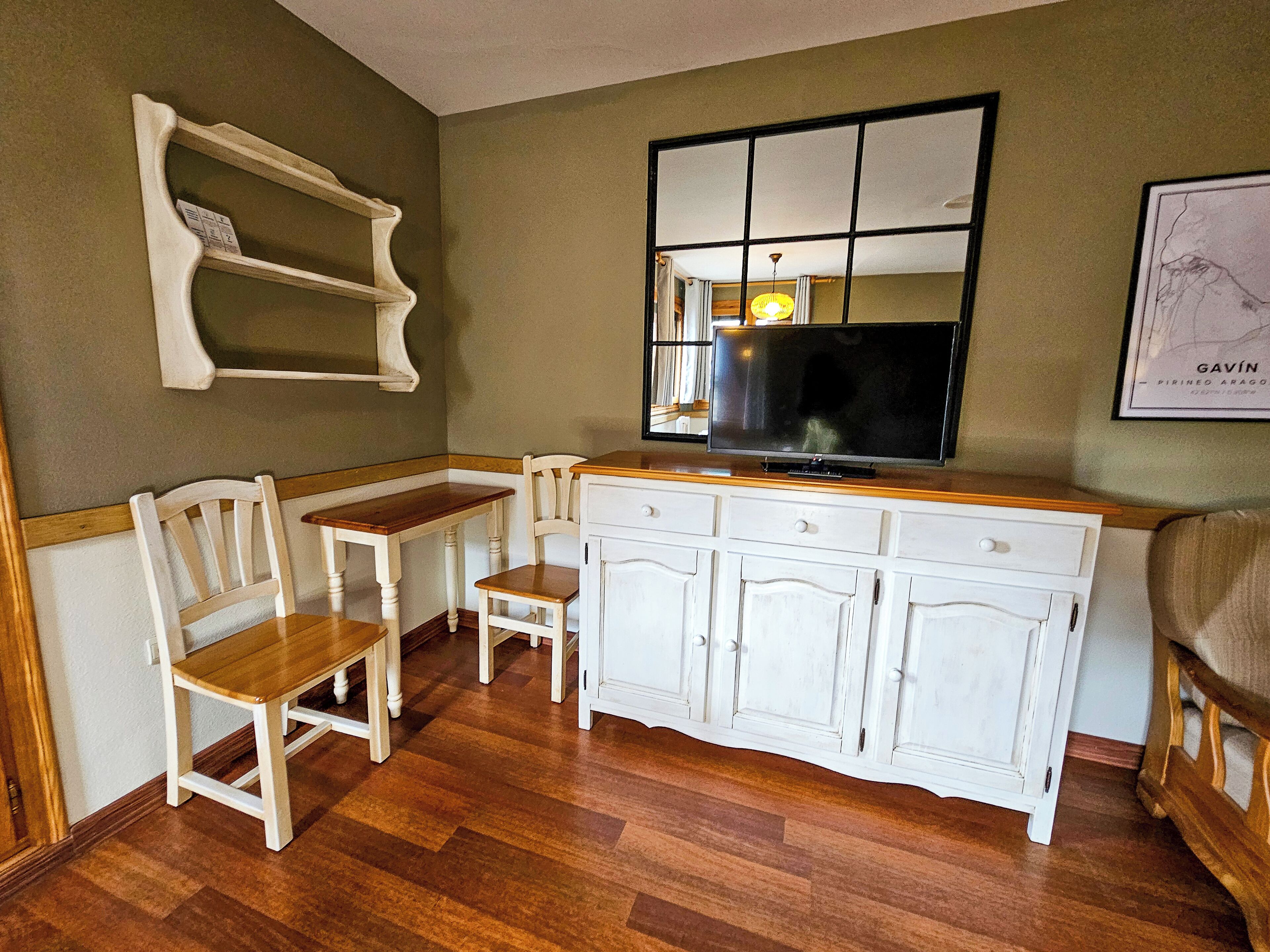 Un espacio habitable pequeño con una mesa de comedor de madera, dos sillas, un armario blanco y un televisor de pantalla plana montado encima.