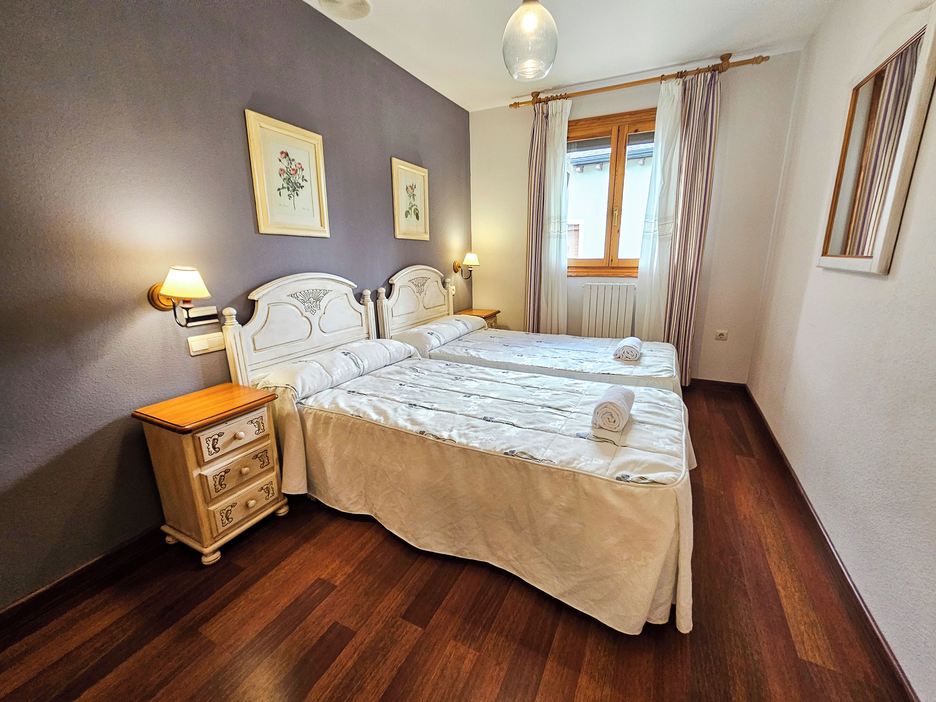 Un dormitorio con dos camas, mesitas de noche de madera, un aparador y una ventana con cortinas.