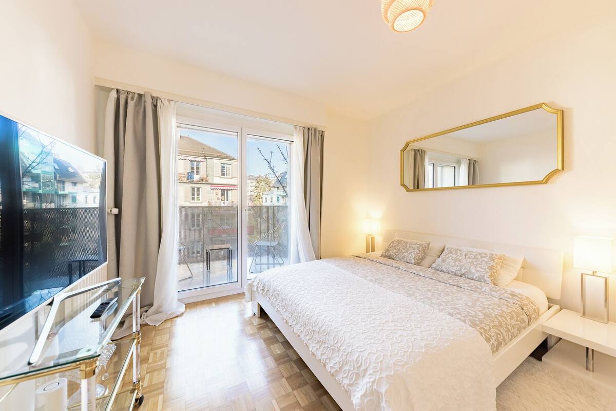 Una habitación de hotel con una cama grande, televisión, una mesa de vidrio y un balcón con vistas a edificios.