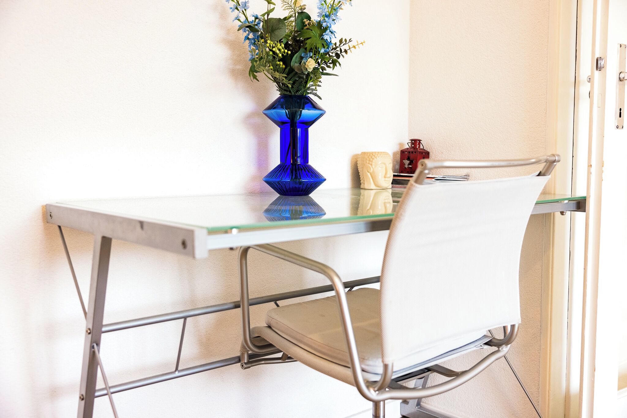 A dining area with a glass table, metal chair, and a blue vase with flowers.
