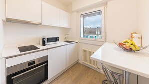 Une cuisine moderne avec des meubles blancs, un four à micro-ondes, un four traditionnel, une fenêtre donnant sur des immeubles et un panier de fruits posé sur le plan de travail.