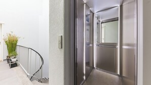 A modern elevator with stainless steel doors and a glass panel, a metal handrail, and a potted plant beside a white wall.