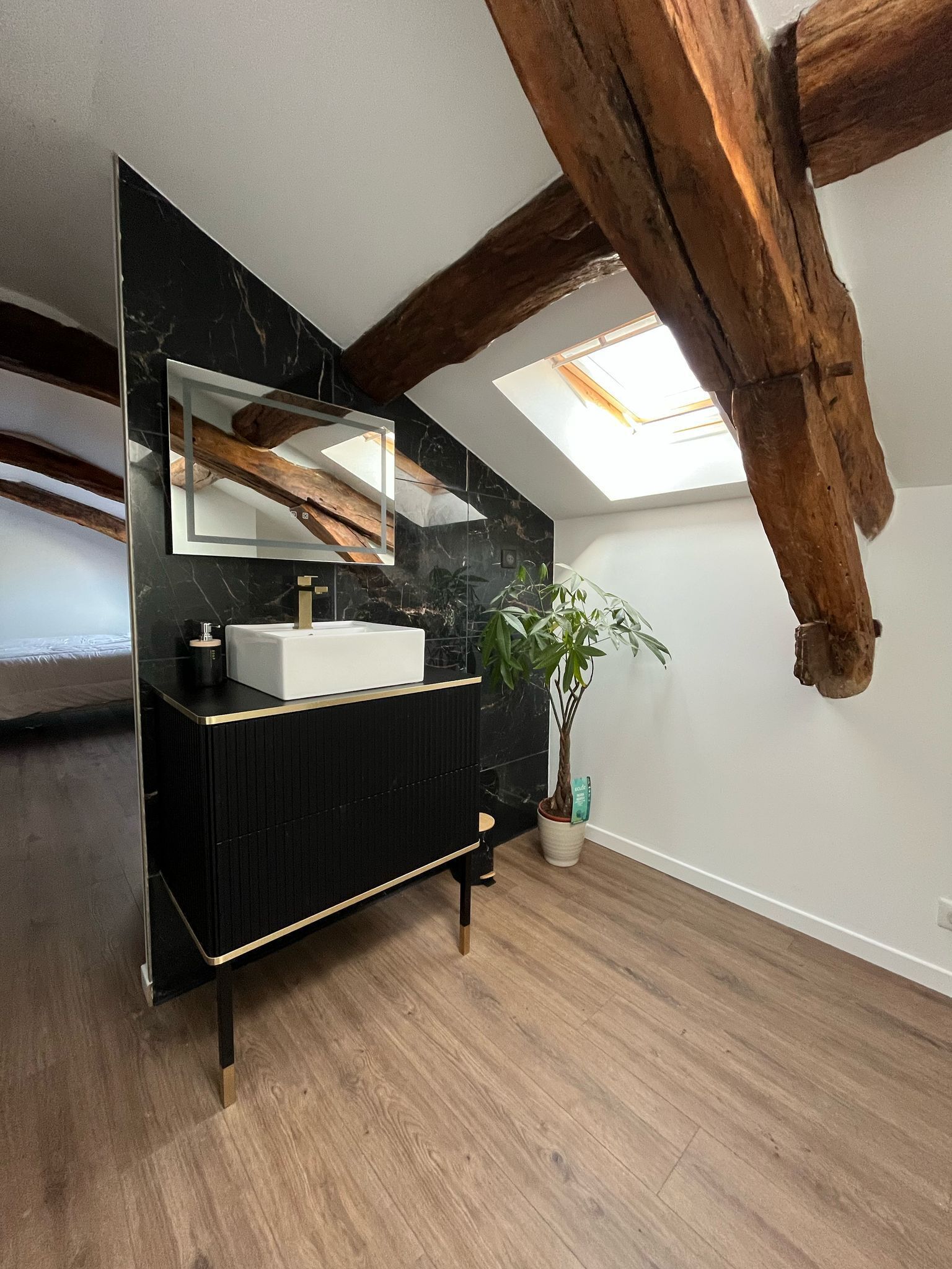 Een moderne badkamer met een zwart wastafelmeubel, een witte wasbak en een grote spiegel. Het plafond heeft zichtbare houten balken en er is een dakraam.