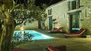 Μια νυχτερινή θέα μιας περιοχής δίπλα στην πισίνα, με ένα πέτρινο κτίριο, μια ξύλινη βεράντα και υπαίθρια καθιστικά.