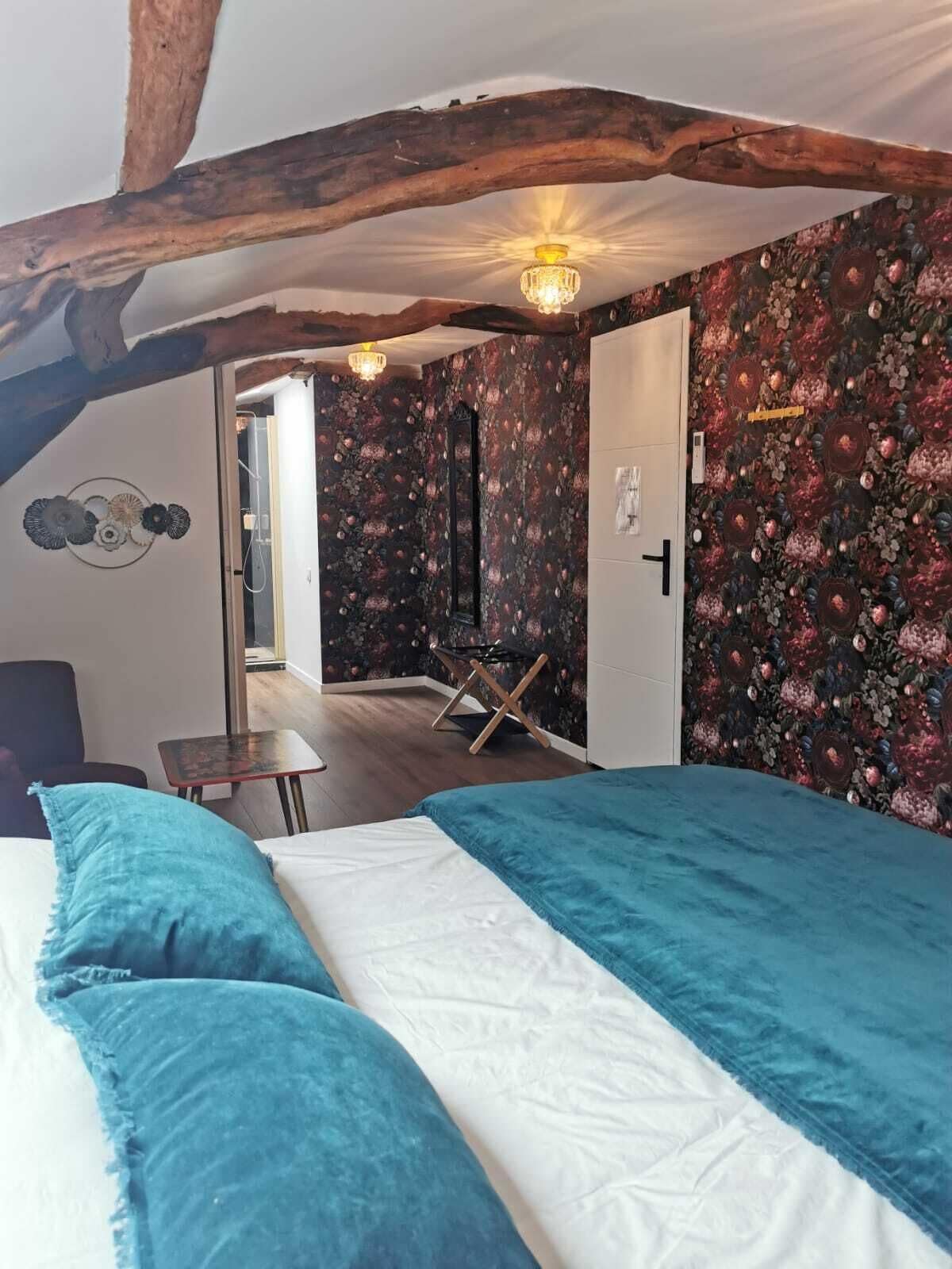 A bedroom with a bed, a wooden ceiling beam, a floral wallpaper, and a door leading to another room.