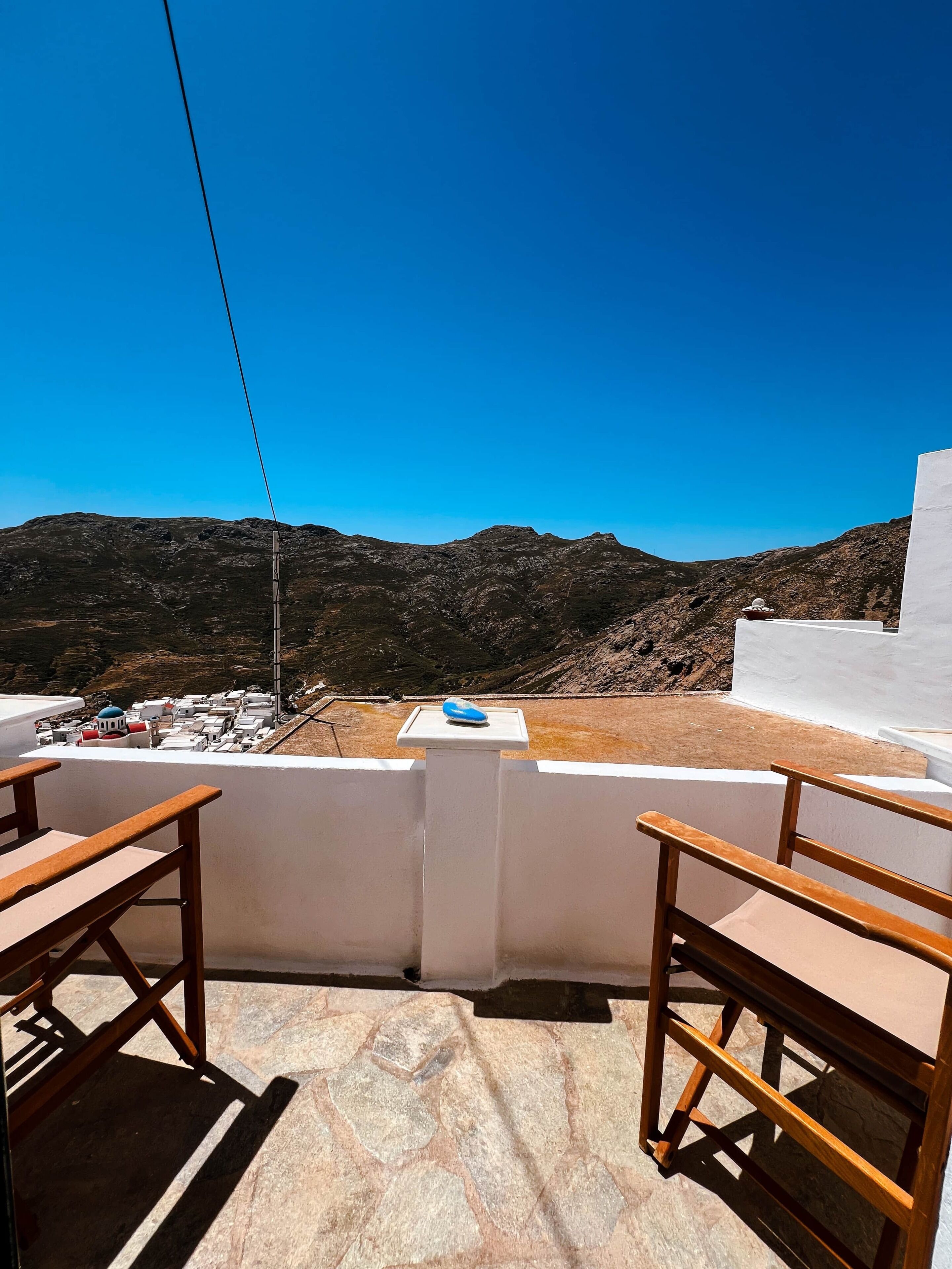 A terrace with a view of a mountainous landscape, a white wall, and two wooden chairs.