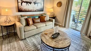 A living room with a sofa, two side tables, a coffee table, a large painting, and a view through a glass door. - Romantic Smoky Mountain Escape-Hot tub-Pool-fireplace (Gatlinburg)