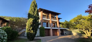 A multi-story building with a brick facade, balconies, and a well-maintained garden with a tall tree and shrubs.